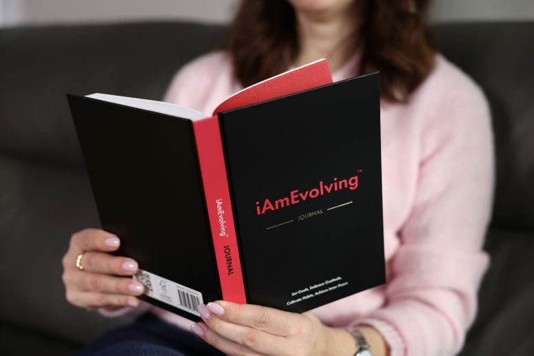 Lady holding a book wiht a black and read cover that says iAmEvolving, Journel. She is sitting down wearing a pink sweater