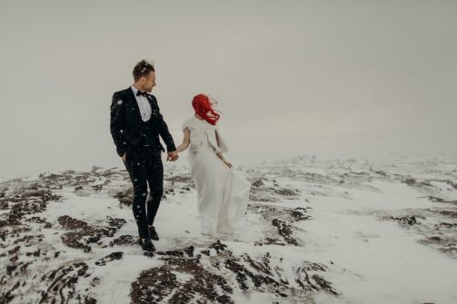 couple getting maqrried in snow, man is in black and white suite, and woman is in white dress with bright red hair.