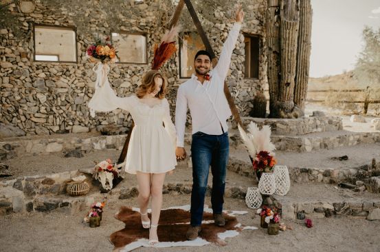 Couple standing in front of simple wedding alter with flowers sparsly around in thrifted jars. They are wearing nontraditional wedding outfits to stick within a budget
