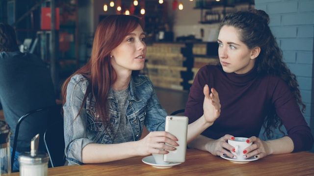 Two sisters arguing in coffee shop one is holding a phone and the other is looking at her. one is a redhead wearing a jean jacked, and the other is brunett in a maroon turtleneck